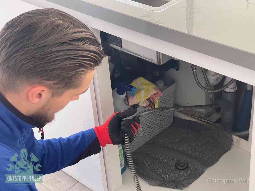 Een monteur voert een flexibele ontstoppingsveer in onder een keukenkastje om een verstopping voorbij de sifon te bereiken in Velp