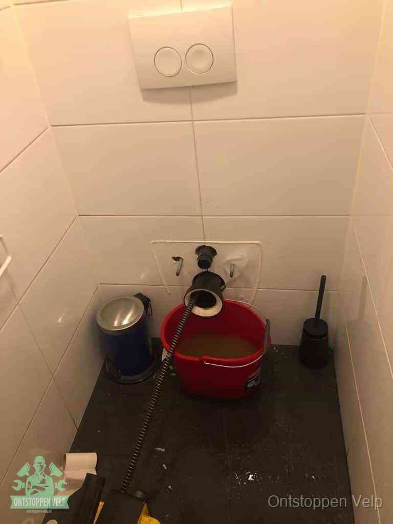 Een ontstopte wand-wc met verwijderde toiletpot waar een ontstoppingsveer in de wanduitgang verdwijnt, het achterhalen waar de verstopping zit in Velp