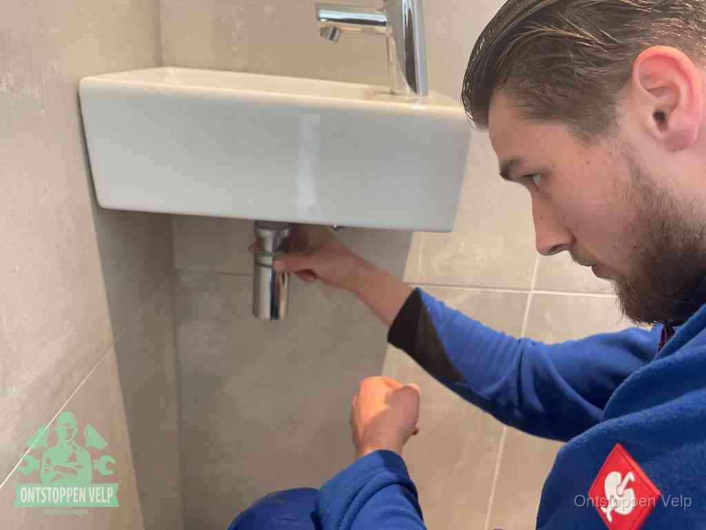 Een monteur draait met de hand de zwanenhalssifon onder een fonteinwasbak in de toiletruimte los om de bron van rioollucht in Velp te vinden