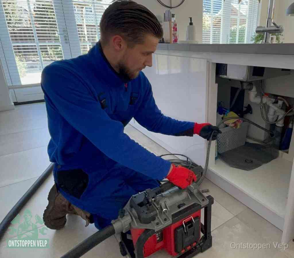 Een monteur met rode handschoenen knielt in een keuken en voert een elektrische ontstoppingsveer in tijdens het professioneel afvoer ontstoppen in Velp