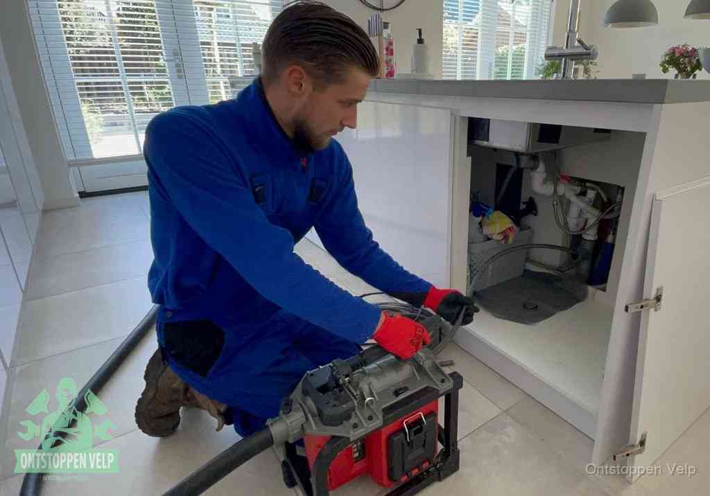 Een monteur voert een professionele elektrische ontstoppingsmachine in de afvoer onder de spoelbak tijdens het gootsteen ontstoppen in Velp